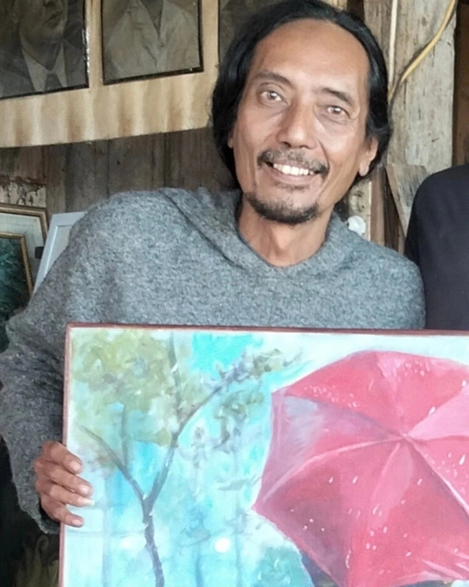 Indonesian artist Heru Japra Pramono smiling and holding his original oil on canvas painting There is Always Love Behind the Red Umbrella in his open barn studio.