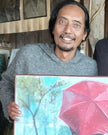 Indonesian artist Heru Japra Pramono smiling and holding his original oil on canvas painting There is Always Love Behind the Red Umbrella in his open barn studio.