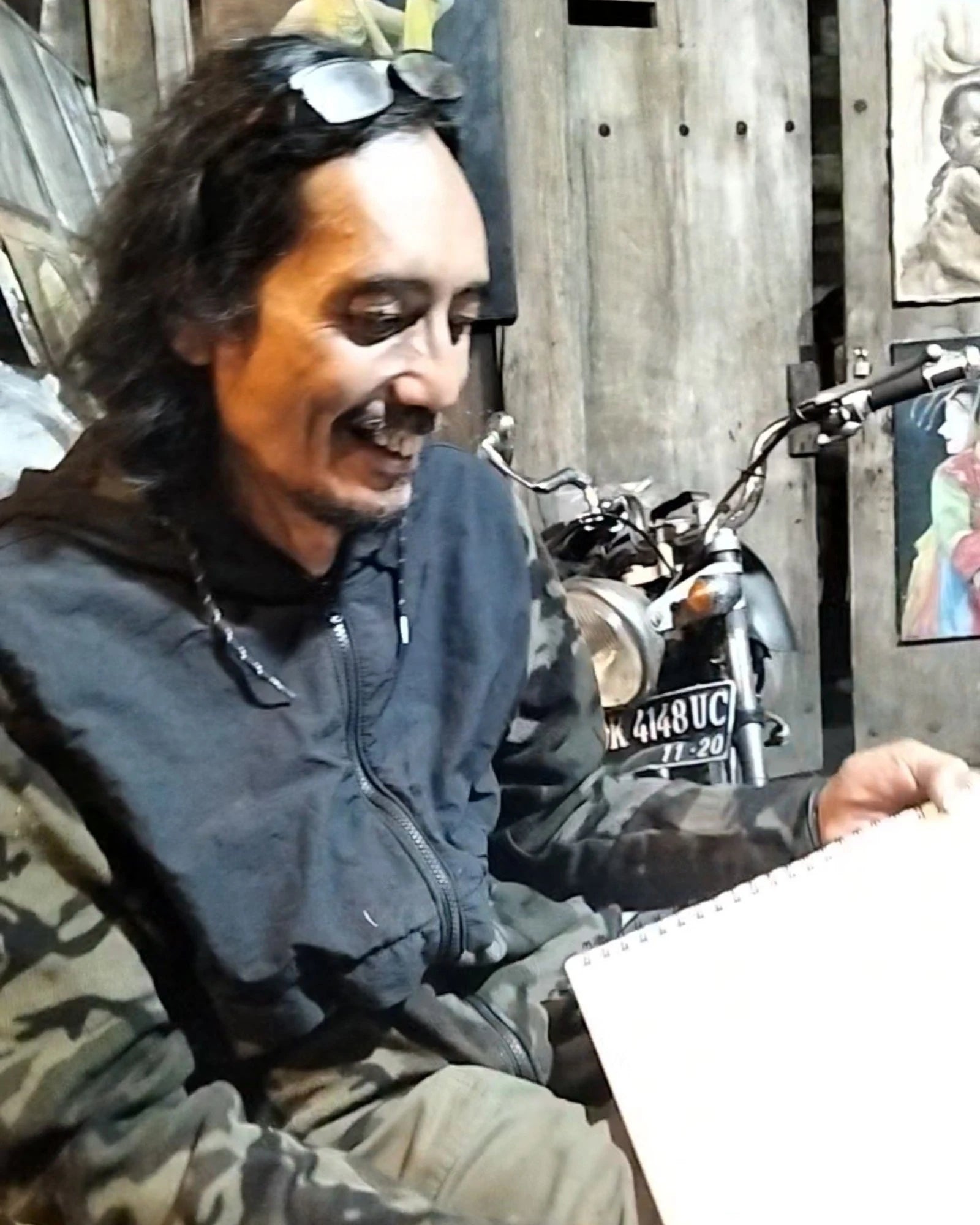 Indonesian artist Heru Japra Pramono smiling and working on a sketch in his open barn roadside art studio in Bali.