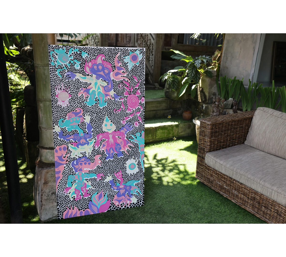 Keep Smiling (2026), an original painting by emerging Balinese artist I Made Ananda Krisna, displayed outdoors on his family’s art gallery property in Bali, beside a traditional stone structure and garden seating — curated and photographed by Senja Living.